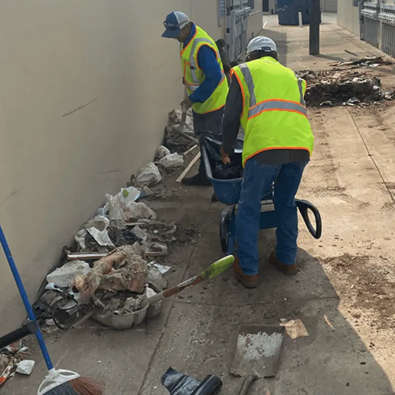 Two CenTex crew in hi-vis vests sweeping debris along a building wall into a wheelbarrow